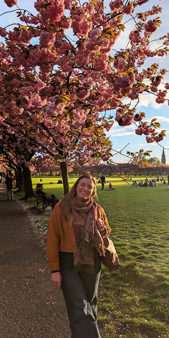 Hannah smiling under a blooming pink cherry blossom tree in The Meadows, Edinburgh.