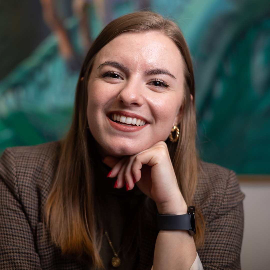 Headshot of the designer, Hannah Graham. She is laughing, resting her chin on her hand, against a blurred green painted background.