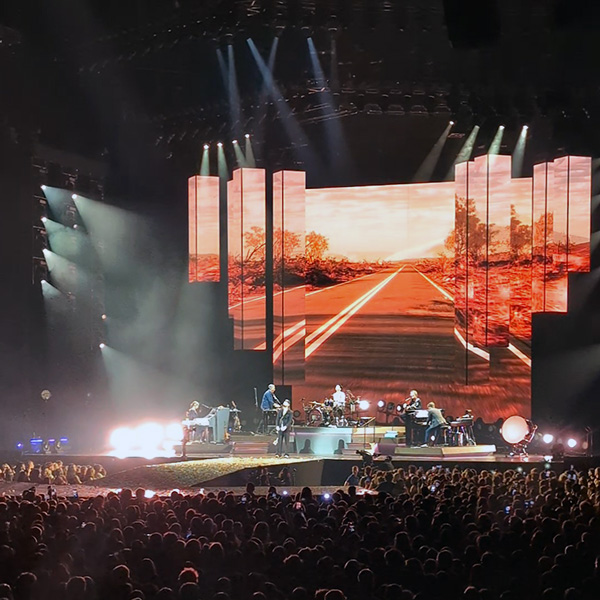 Concert stage with The Lumineers performing in front of large screens showing a sunset road, lit by white stage lights.