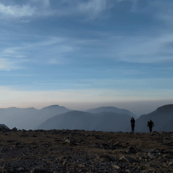 Two hikers silhouetted against a hazy blue mountain range at dusk on a rocky summit.