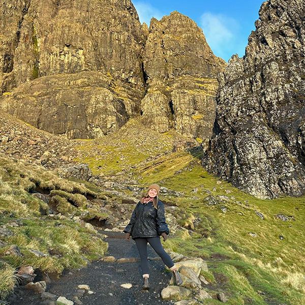 Hannah smiling on a rocky hiking trail beneath the jagged rock formations of the Old Man of Storr, Skye, Scotland.